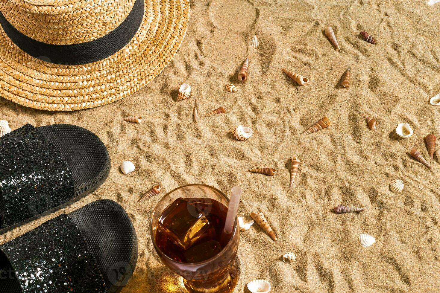 été tenue de plage, retourner flops, chapeau, du froid boisson dans une verre et coquillages sur le sable plage. photo