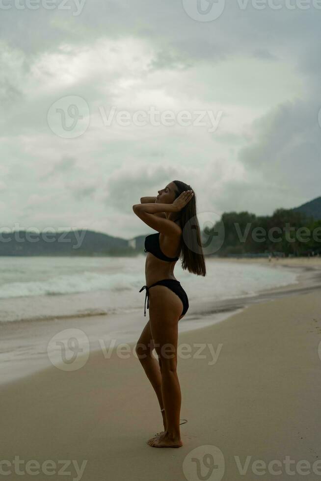 magnifique brunette femme avec parfait figure posant sur tropical plage. portant élégant noir maillots de bain. photo