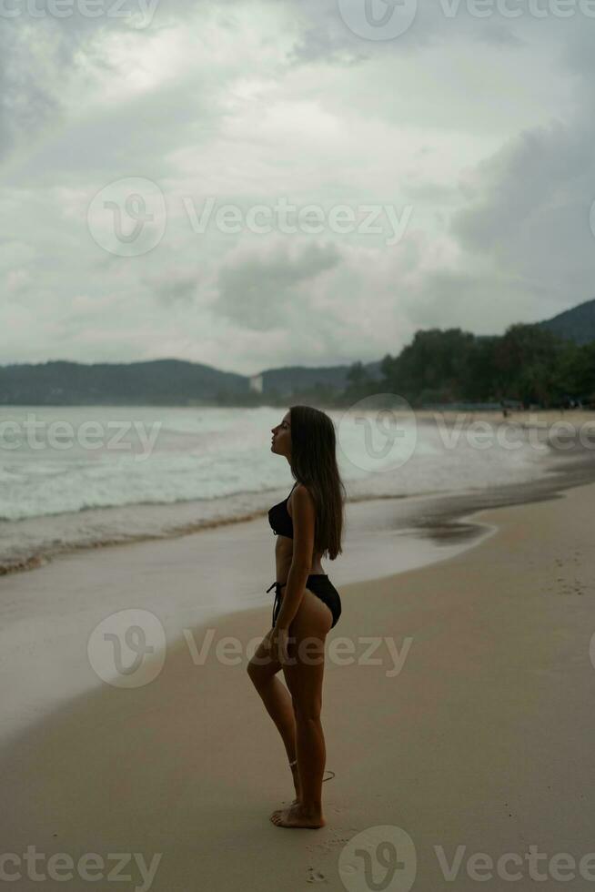 magnifique brunette femme avec parfait figure posant sur tropical plage. portant élégant noir maillots de bain. photo