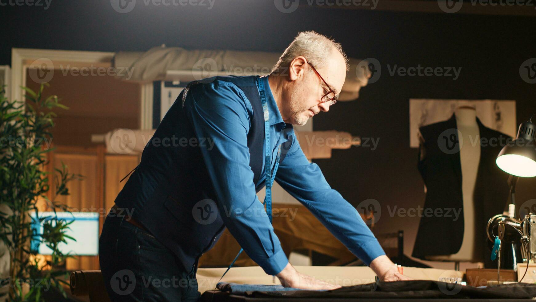 Sénior tailleur dans luxe atelier mesure pièce de en tissu avec mètre à adaptation poste de travail. vieux designer en cours d'analyse croquis à travail sur Vêtements doubler, Douane fabriqué concept. ordinateur de poche tir. photo