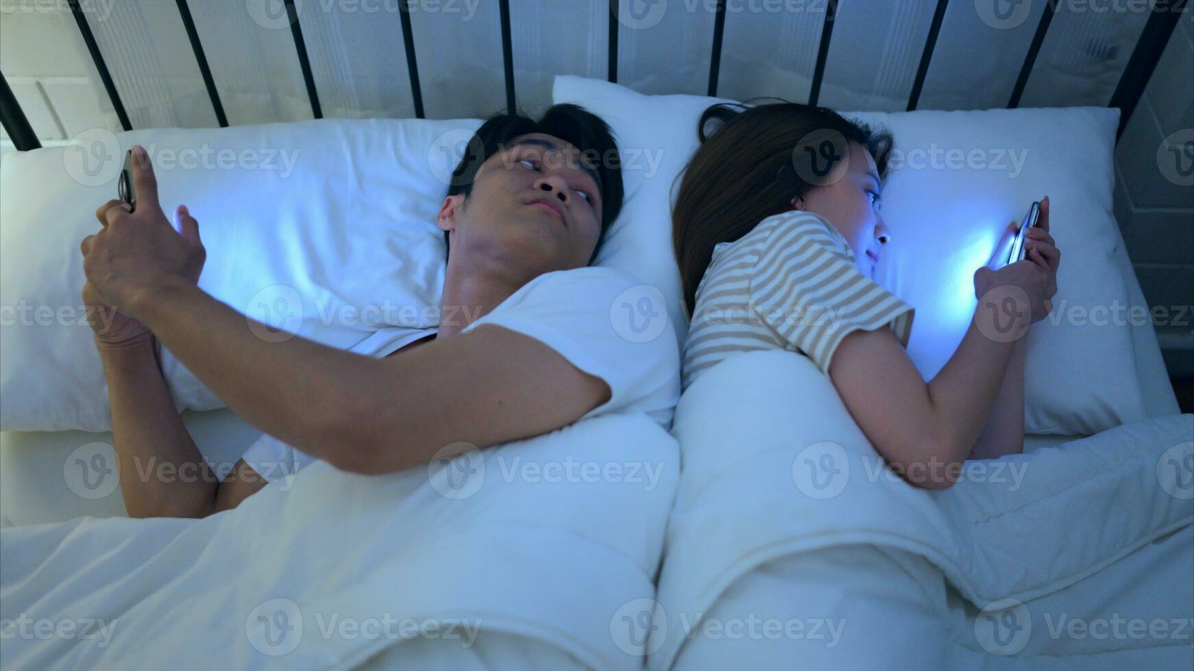 Jeune couple en train de dormir sur le lit dans le chambre à nuit temps avec leur dos à chaque autre et en jouant sur leur Téléphone (s, ignorer chaque autre. photo