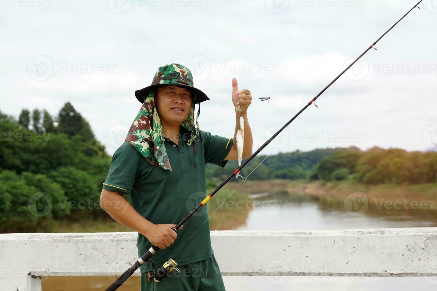 un asiatique homme en portant une pêche barre et le sien capture des stands sur une pont plus de une rivière dans le village où il est une ordinaire pêcheur sur vacances. et c'est une amusement et relaxant Extérieur activité. photo