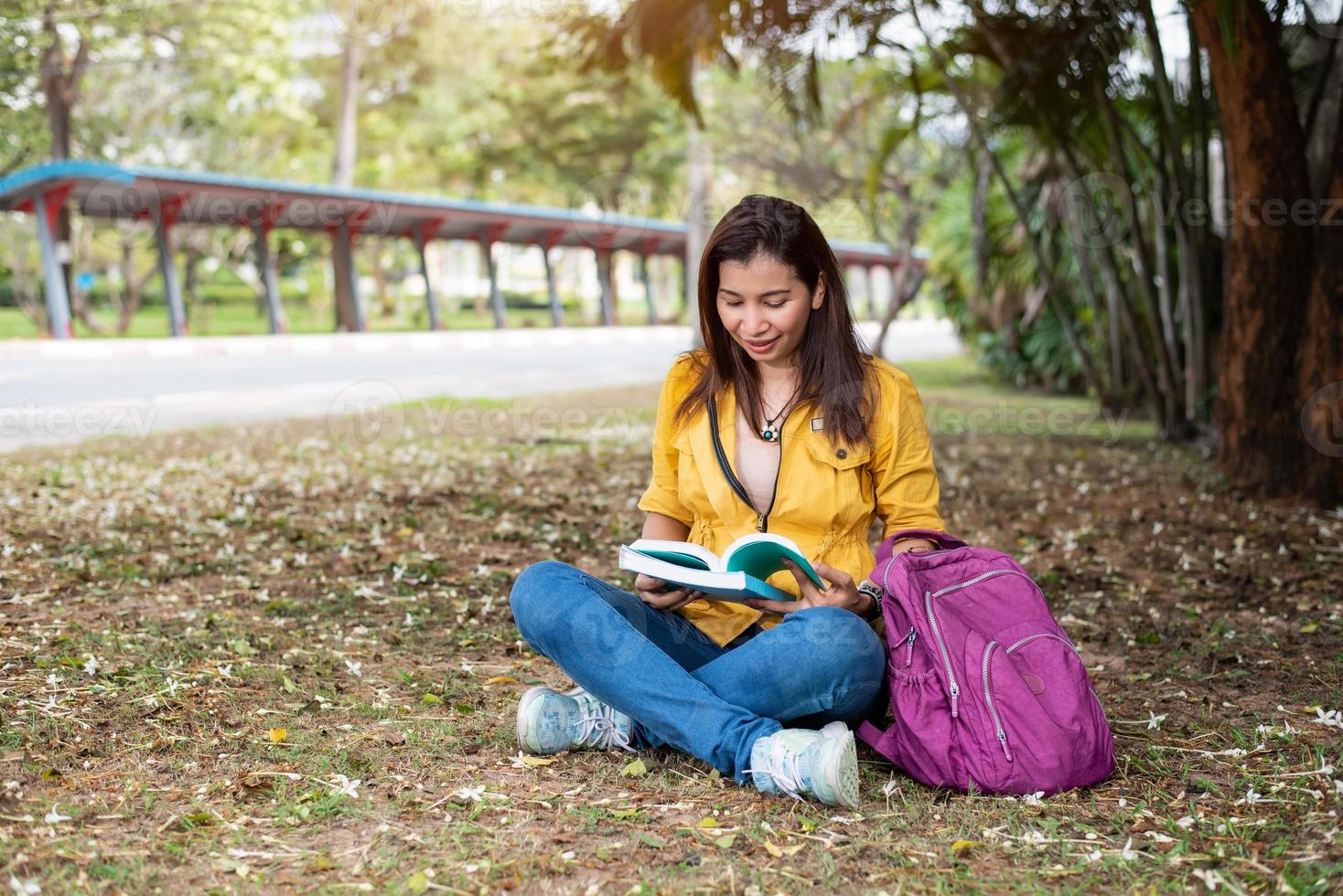 Heureuse femme asiatique assise et lisant des livres dans le parc universitaire photo