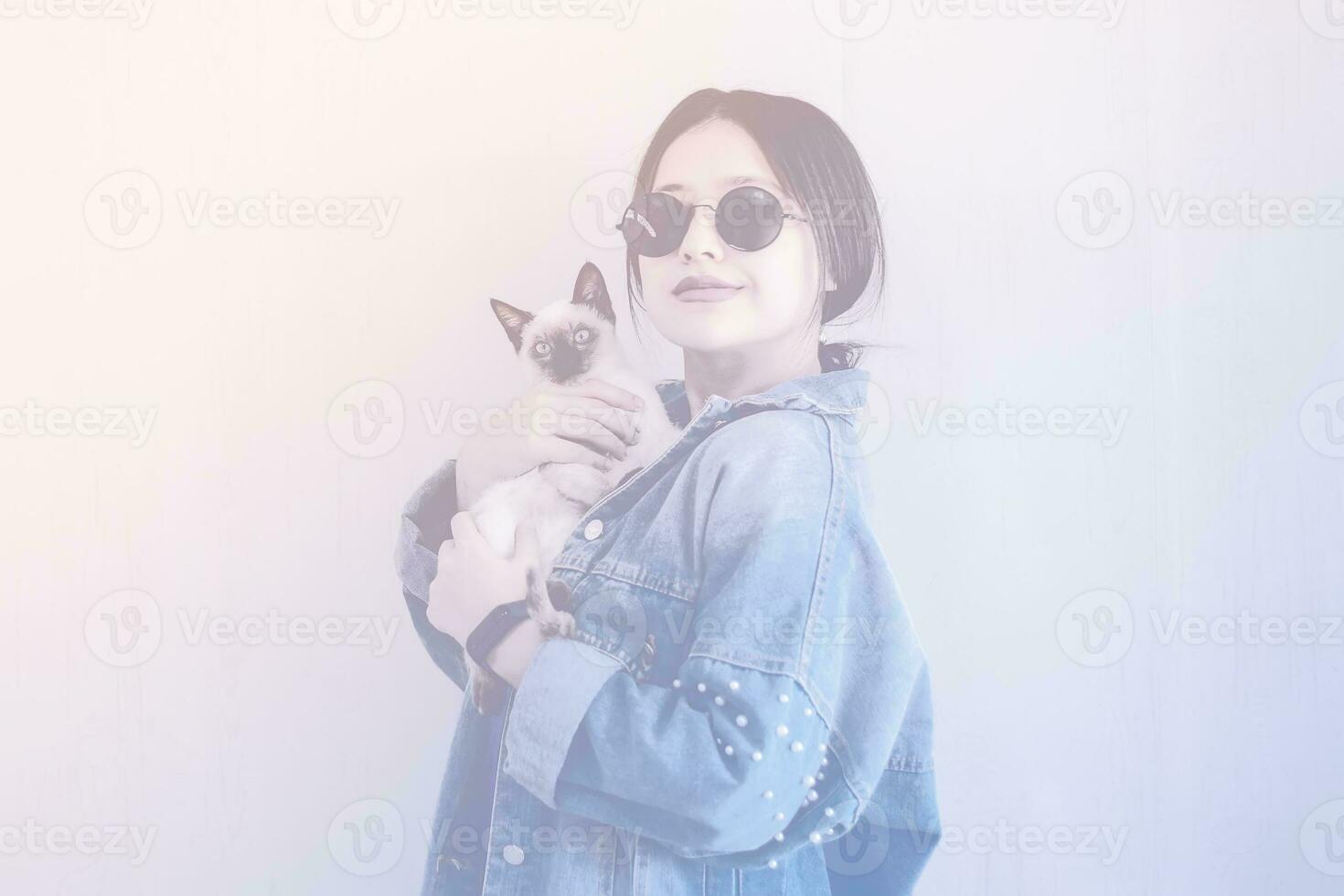 Jeune attrayant femme étreindre chatte chat dans mains. mignonne et glamour fille dans branché des lunettes de soleil posant avec sa Siamois chat photo