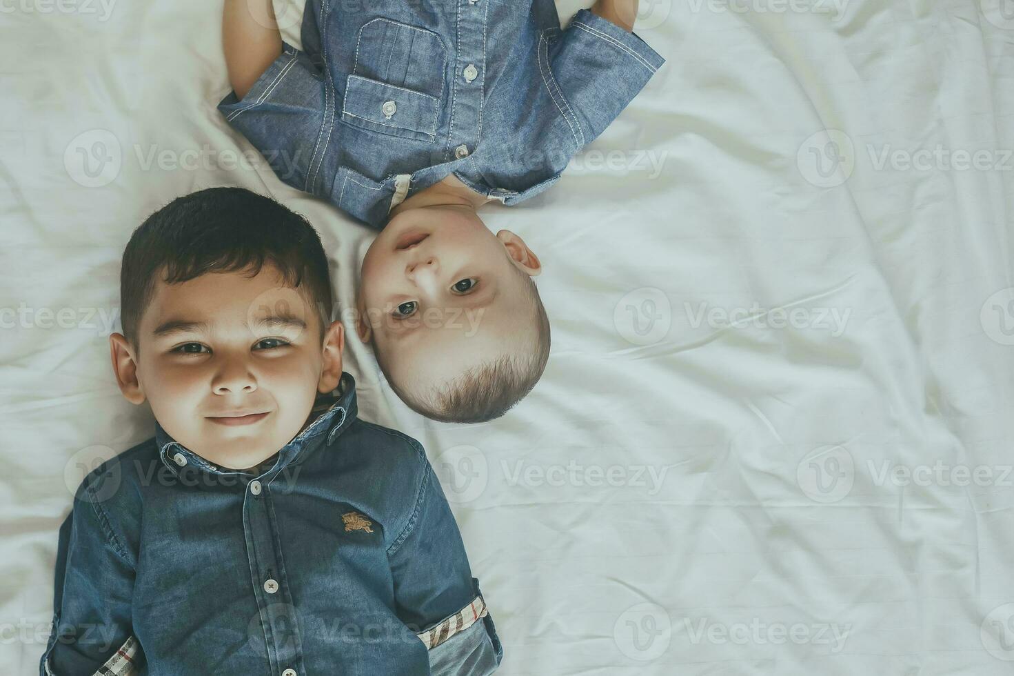 content enfance concept. content frères portrait. 6 ans garçon et 6 mois vieux bébé garçon ayant amusement. deux peu des gamins souriant à le caméra. mignonne peu frères mensonge sur lit ensemble photo