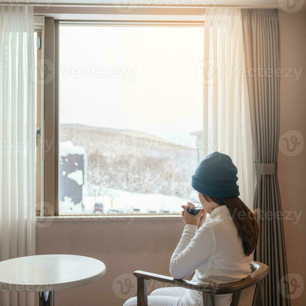 Jeune femme dans chandail avec tasse de café à la recherche par le fenêtre dans hiver saison, content femelle profiter chute de neige Extérieur vue à appartement ou Accueil dans le Matin. veille et relaxant concept photo