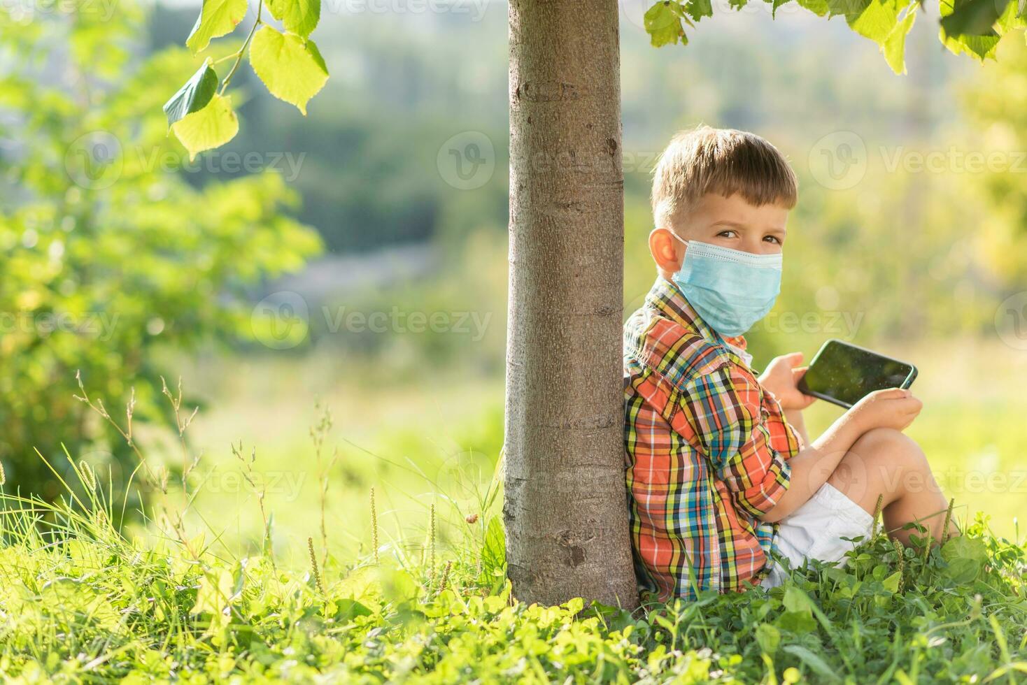 une enfant dans une médical masque est assis sur le herbe et regards dans le téléphone les dessins animés dans le été à le coucher du soleil. enfant avec une mobile téléphone dans le sien mains. la prévention contre coronavirus covid-19 pendant une pandémie photo