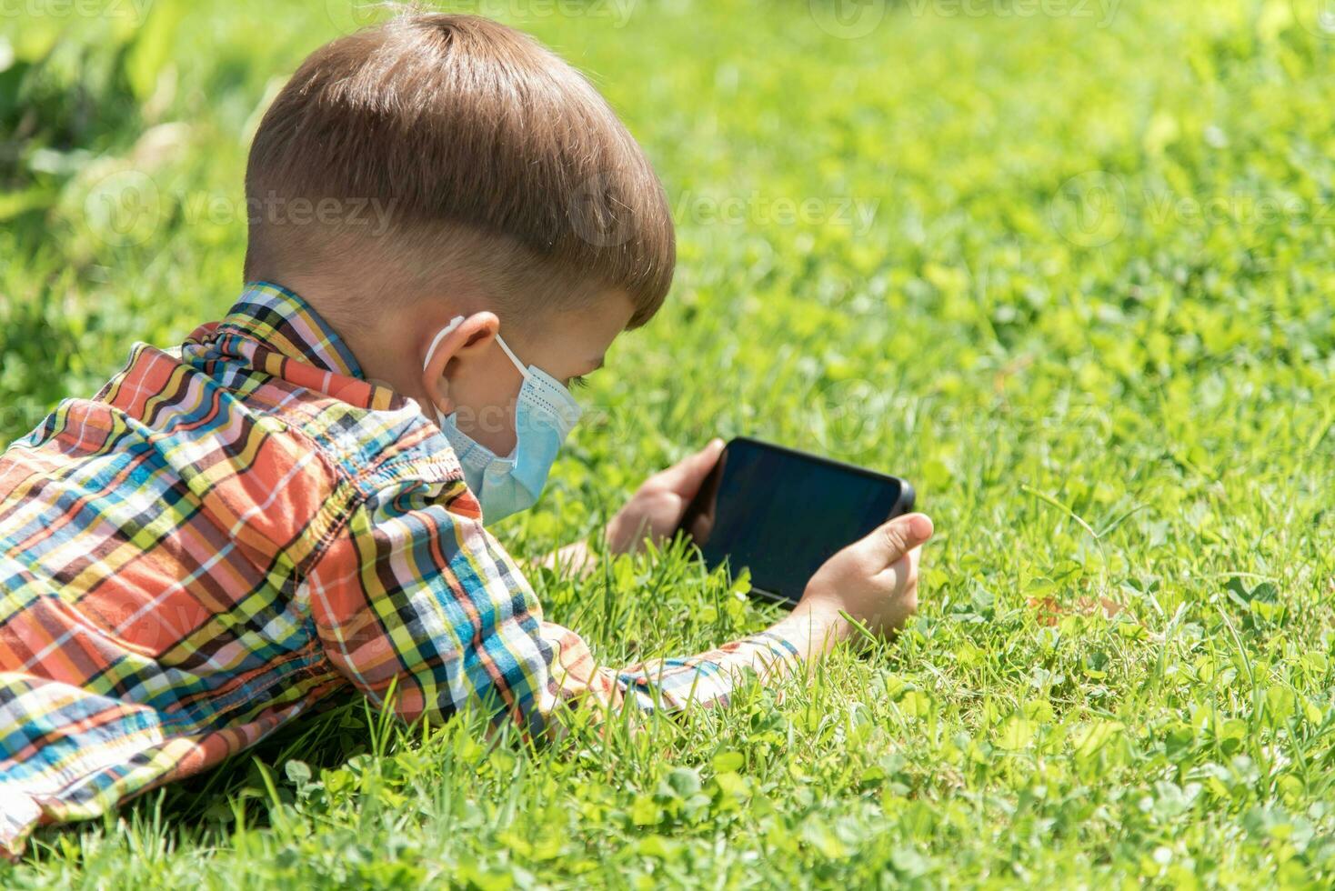 une enfant dans une médical masque mensonges sur le herbe et regards dans le téléphone les dessins animés dans le été à le coucher du soleil. enfant avec une mobile téléphone dans le sien mains. la prévention contre coronavirus covid-19 pendant une pandémie photo