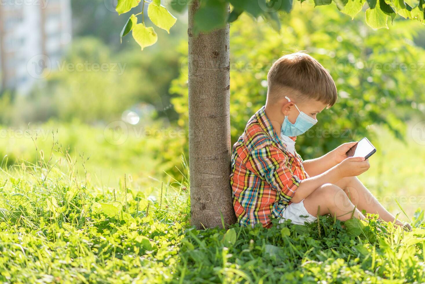 une enfant dans une médical masque est assis sur le herbe et regards dans le téléphone les dessins animés dans le été à le coucher du soleil. enfant avec une mobile téléphone dans le sien mains. la prévention contre coronavirus covid-19 pendant une pandémie photo