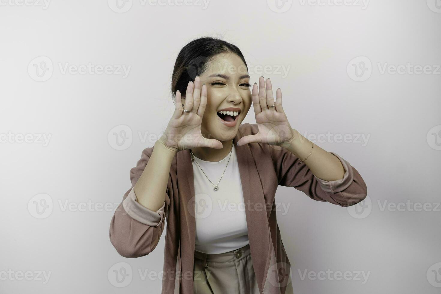 une Jeune magnifique asiatique femme employé portant cardigan est en criant et en hurlant bruyant avec une main sur sa bouche, isolé par blanc Contexte. photo