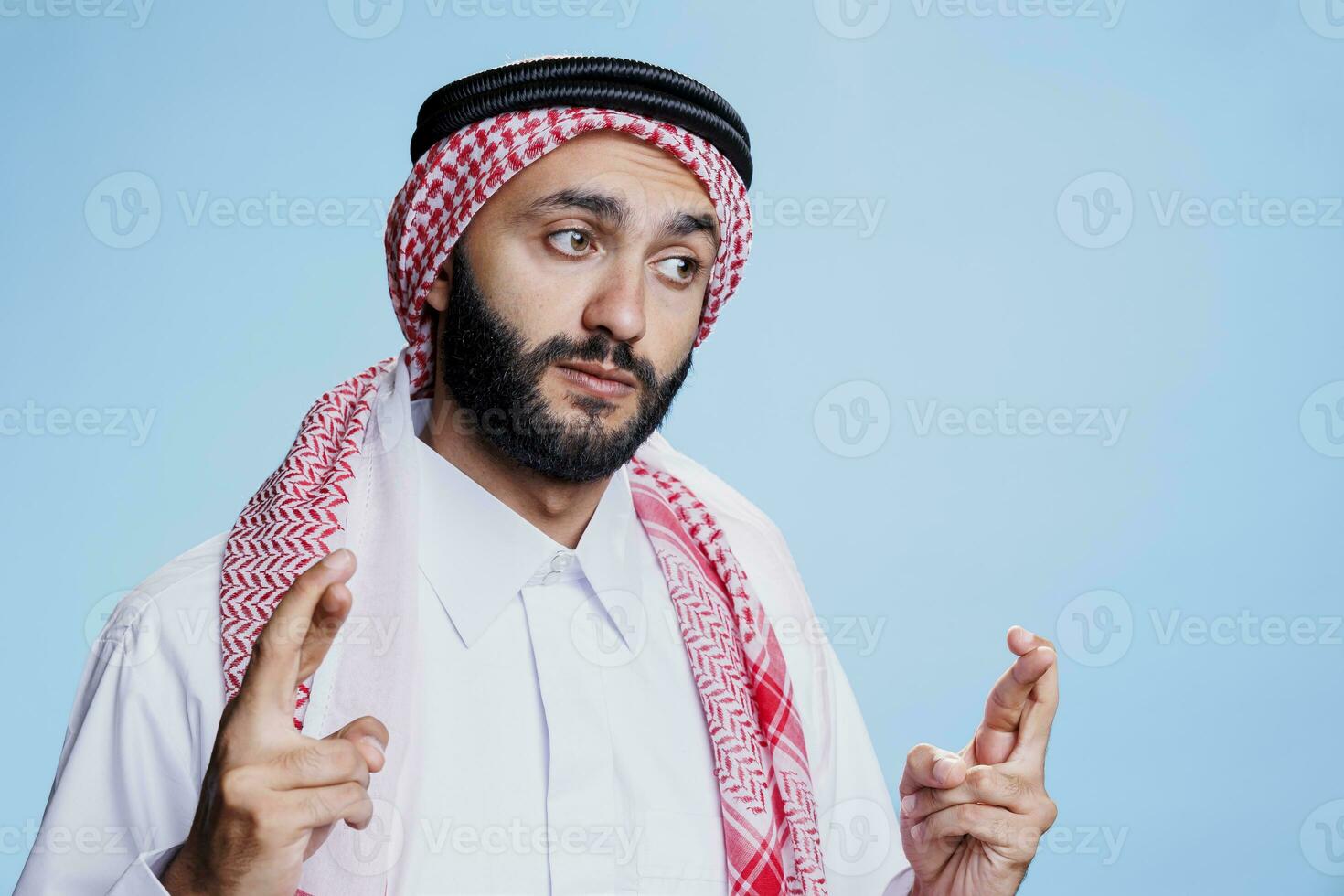 arabe homme portant traditionnel thobe et foulard traversée les doigts pour bien chance. la personne habillé dans islamique culturel vêtements montrant superstitieux geste, plaidoirie pour Succès photo