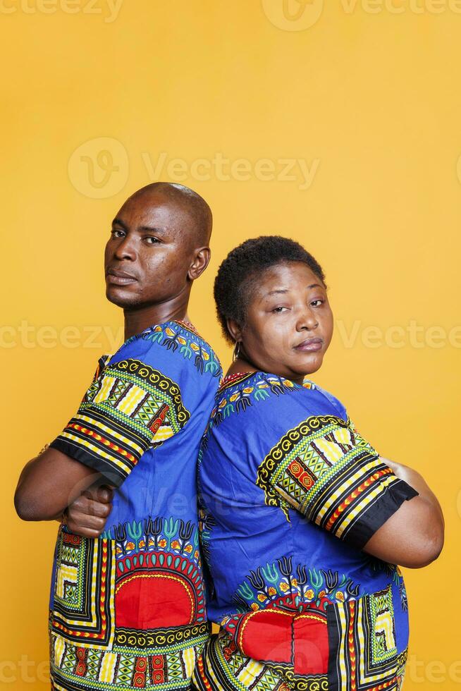 sérieux noir couple habillé dans ethnique vêtements posant avec bras franchi studio portrait. sur de soi homme et femme permanent retour à retour avec plié mains et à la recherche à caméra photo