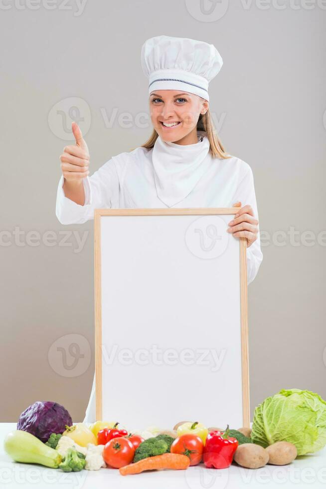 de bonne humeur femelle chef est montrant pouce en haut tandis que en portant tableau blanc avec bouquet de des légumes sur le tableau. photo