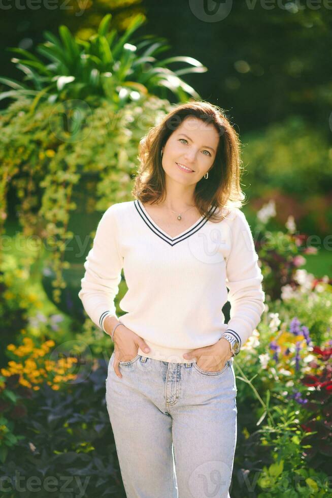 Extérieur portrait de content magnifique 40 - 45 année vieux femme, portant blanc arrêtez-vous, posant dans parc sur une très ensoleillé jour, en bonne santé mode de vie photo