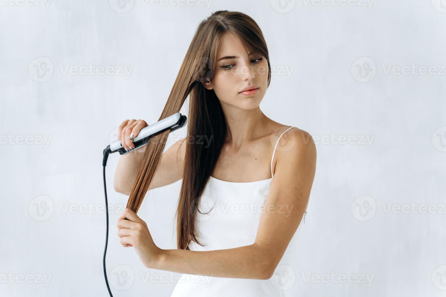 adorable jeune femme lissant ses cheveux avec un fer plat et regardant de côté avec des émotions calmes. mur blanc à l'arrière-plan. concept de cheveux de femme photo