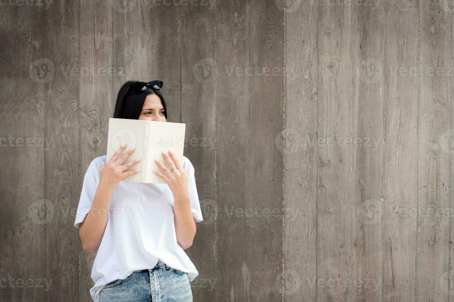 contre le Contexte de une gris mur, une Jeune étudiant timidement couvert sa visage avec une livre photo