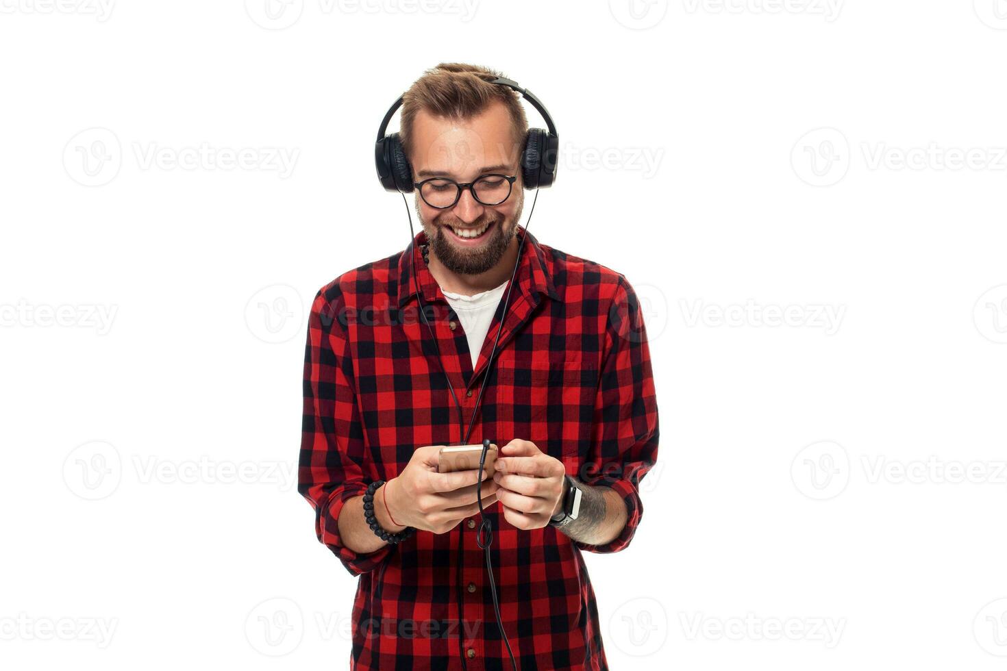 Jeune branché homme dans à carreaux chemise et des lunettes portant écouteurs à la recherche content sur blanc Contexte. photo