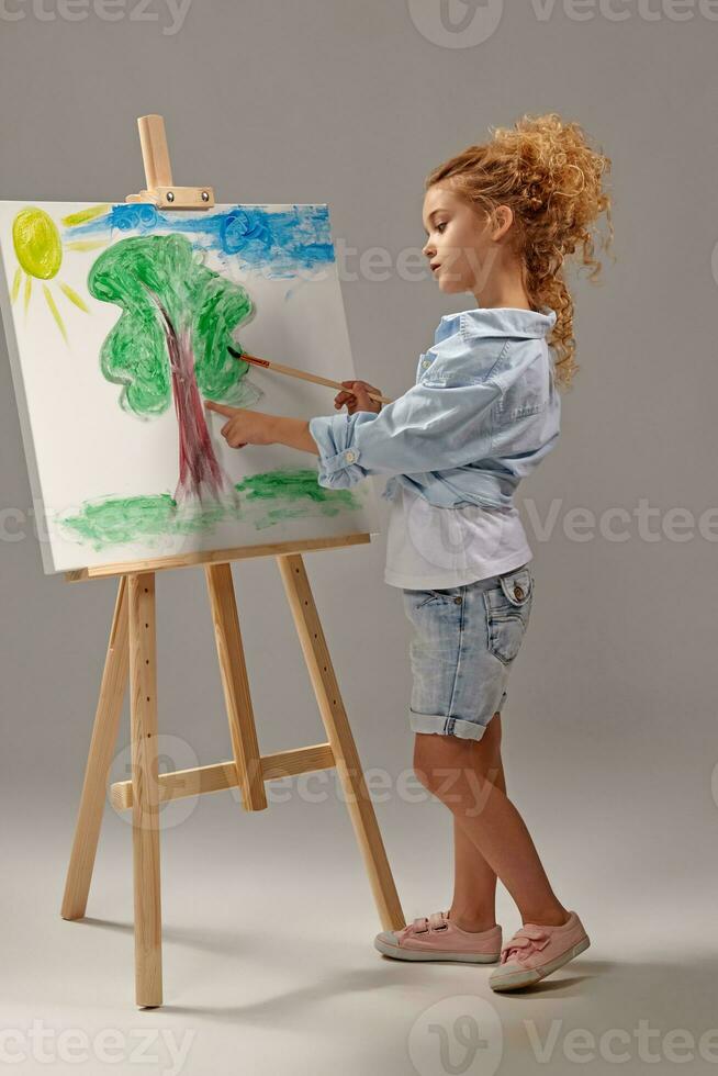 charmant école fille est La peinture avec une aquarelle brosse sur un chevalet, permanent sur une gris Contexte. photo