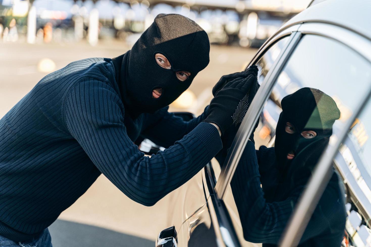 fais attention. homme vêtu de noir avec une cagoule sur la tête en regardant la vitre de la voiture avant le vol. voleur de voiture, concept de vol de voiture photo