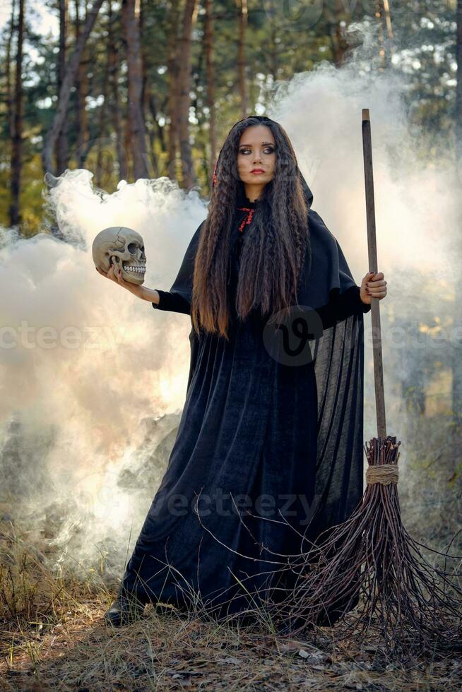 sorcière dans noir, longue robe avec cap et capot. posant dans fumée pin forêt. en portant une crâne et une balai. sorts, la magie et la sorcellerie. plein longueur. photo