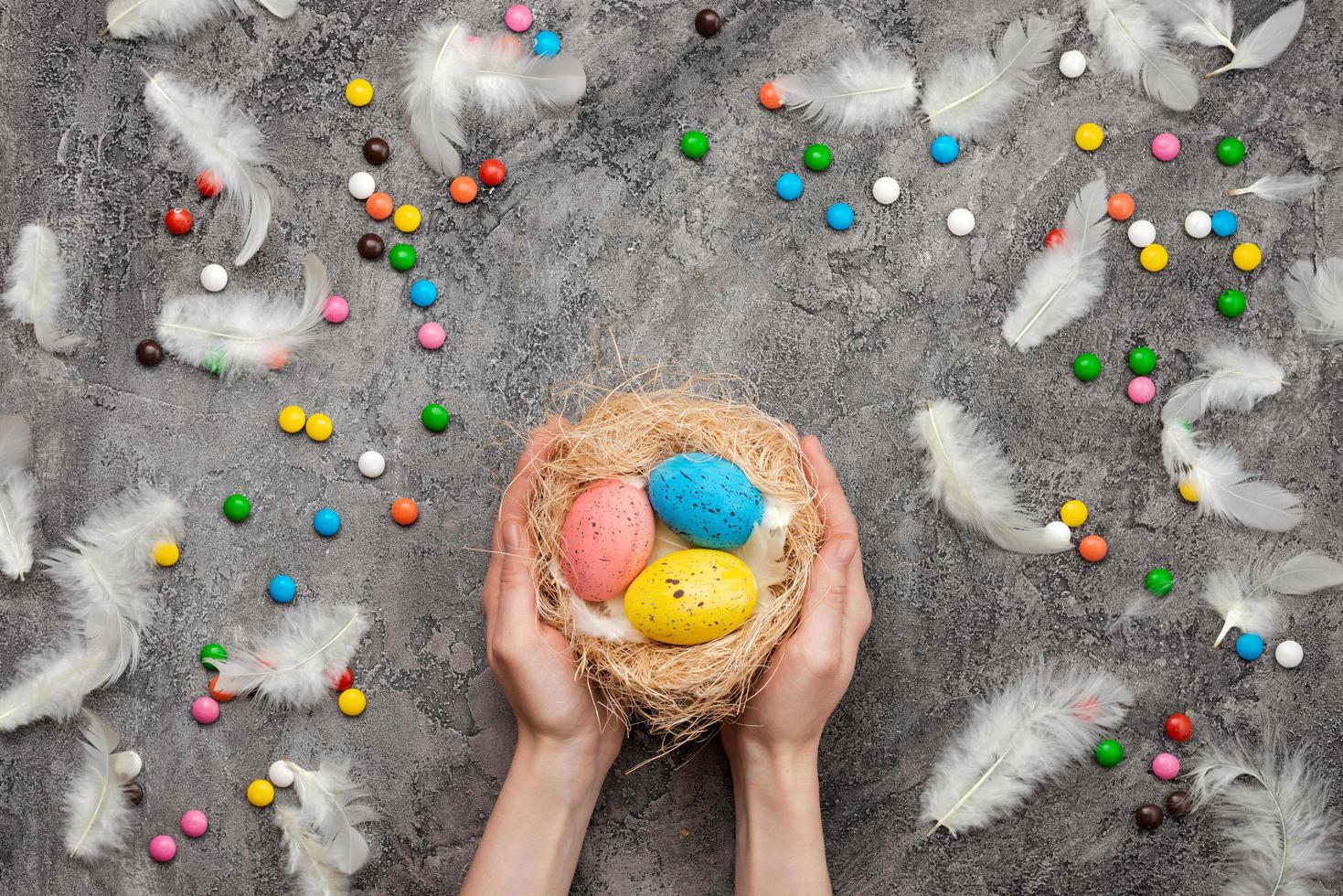 fond de pâques, mains féminines tenant un nid avec des œufs multicolores. bonbons et plumes éparpillés photo