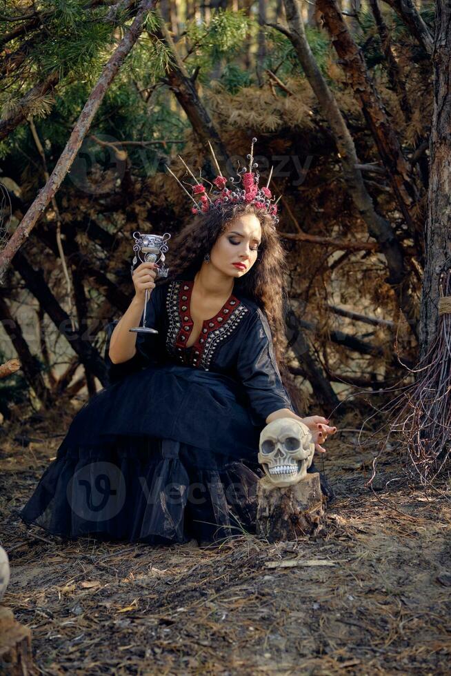 sorcière dans noir, longue robe, avec rouge couronne dans sa longue cheveux. posant avec balai et crâne dans pin forêt. sorts, la magie et la sorcellerie. plein longueur. photo