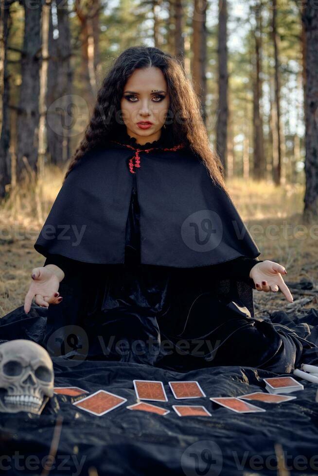 sorcière dans noir robe avec cap et capot. posant dans pin forêt. séance sur foncé couverture avec voyance cartes et crânes sur il. plein longueur. photo