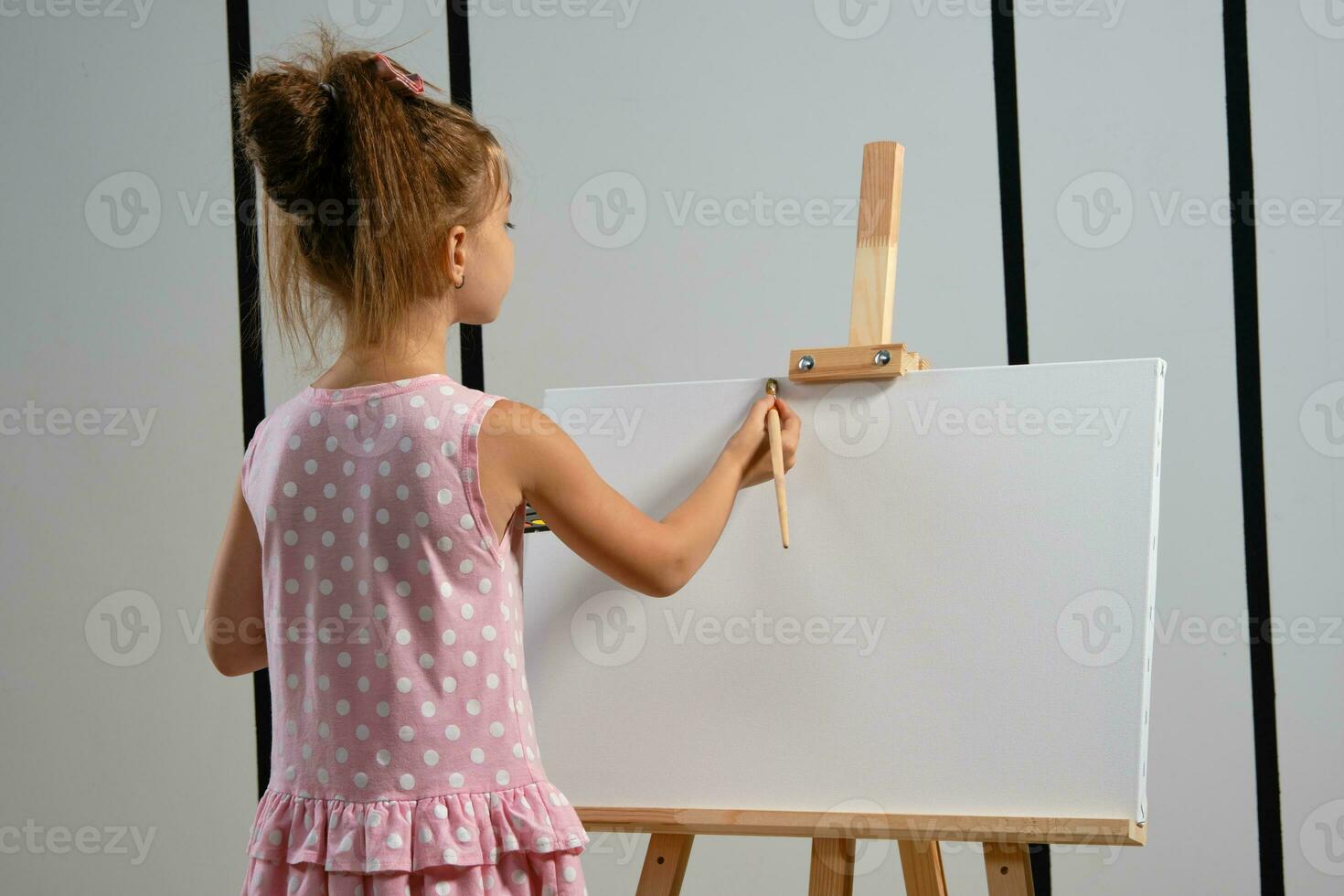 peu fille artiste dans une rose robe est permanent derrière chevalet et La peinture avec brosse sur Toile à art studio avec blanc des murs. moyen fermer tir. photo