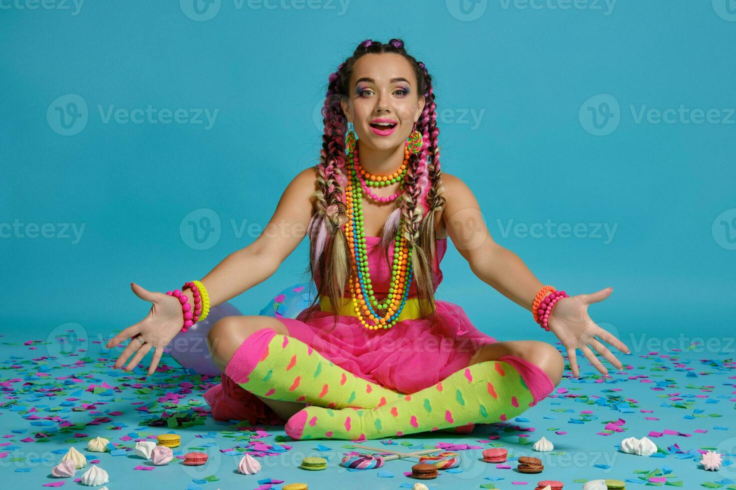 charmant fille avec une multicolore tresses coiffure et brillant se maquiller, posant dans studio avec air des ballons et confettis contre une bleu Contexte. photo