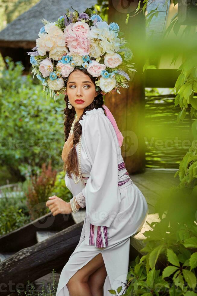 brunette fille dans une blanc ukrainien authentique nationale costume et une couronne de fleurs est posant contre une vert cour. fermer. photo