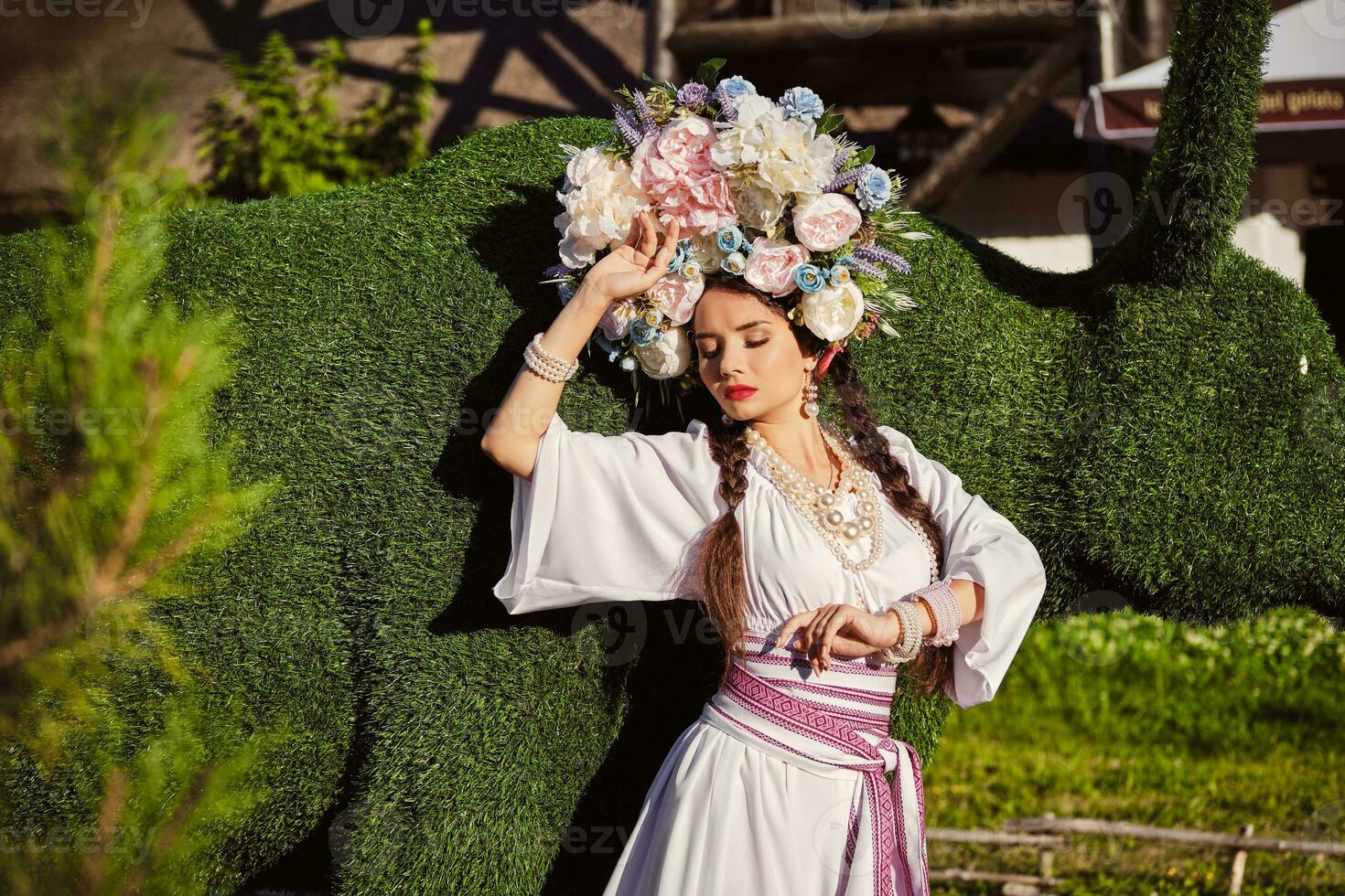 brunette fille dans une blanc ukrainien authentique nationale costume et une couronne de fleurs est posant contre une vert bœuf sculpté de une buisson. photo
