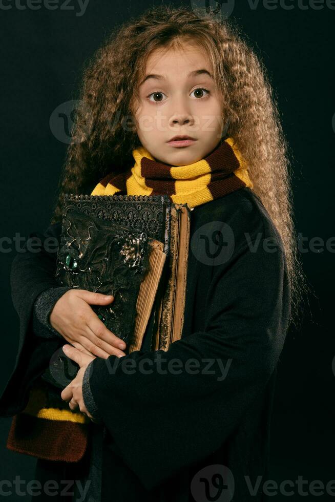 portrait de une peu sorcière fille avec longue marron cheveux habillé dans foncé manteau, en portant la magie livres dans sa main, posant sur noir studio Contexte. photo