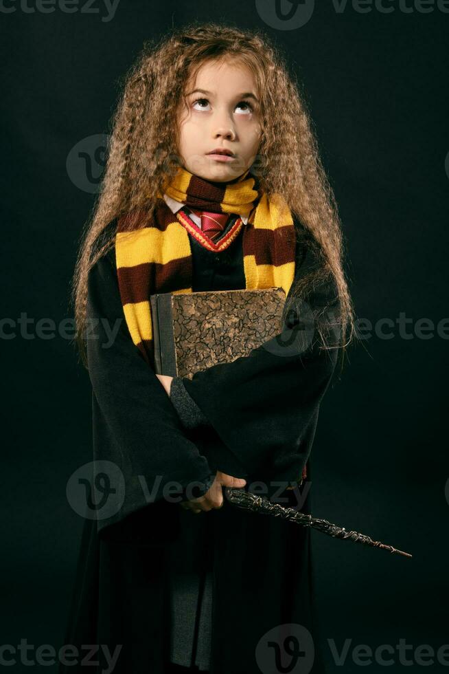 portrait de une peu sorcière fille avec longue marron cheveux habillé dans foncé manteau, en portant la magie baguette magique dans sa main, posant sur noir studio Contexte. photo