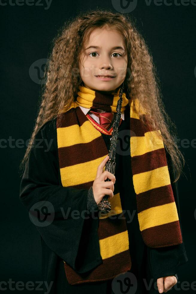 portrait de une peu sorcière fille avec longue marron cheveux habillé dans foncé manteau, en portant la magie baguette magique dans sa main, posant sur noir studio Contexte. photo