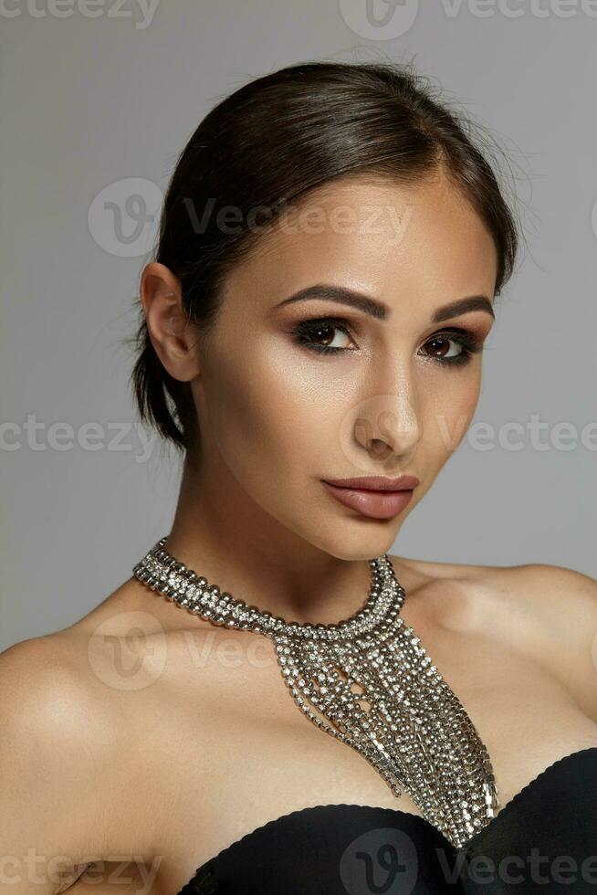 proche en haut portrait de une brunette modèle fille avec professionnel soir se maquiller, portant une noir soutien-gorge et collier, posant sur gris Contexte. photo