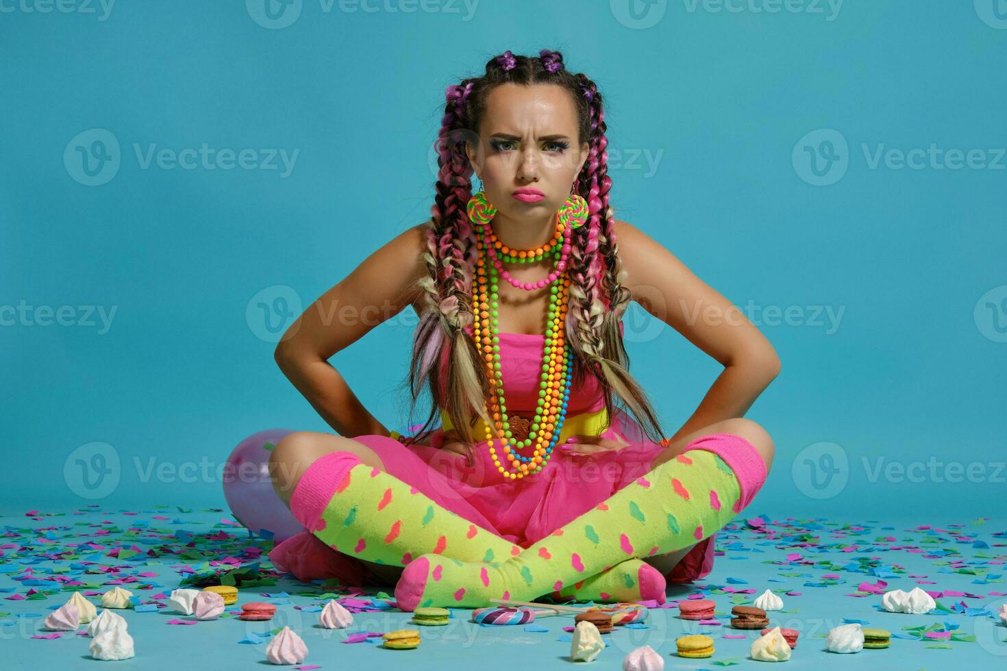 charmant fille avec une multicolore tresses coiffure et brillant se maquiller, posant dans studio avec air des ballons et confettis contre une bleu Contexte. photo