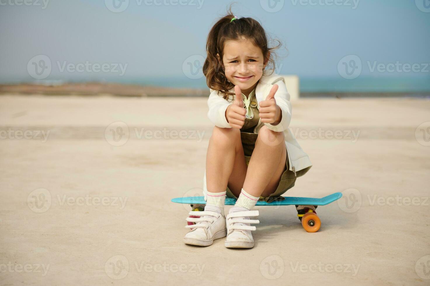 caucasien élégant peu fille enfant séance sur une planche à roulette sur mer promenade, feuilleter en haut et souriant à la recherche à caméra photo