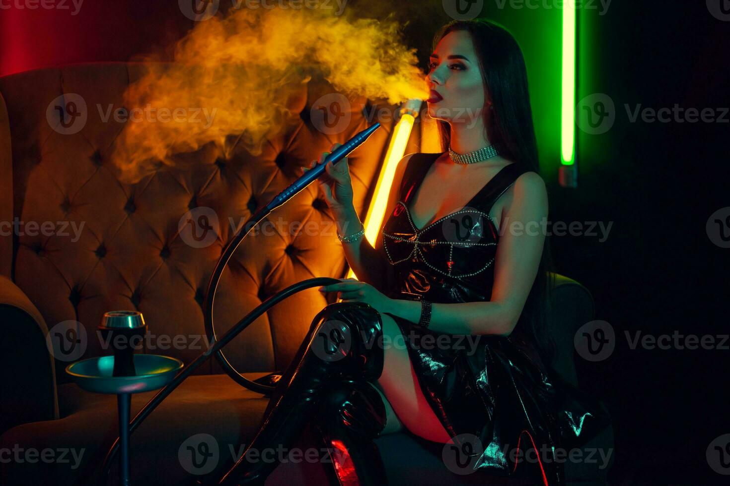 sexy brunette modèle est fumeur une narguilé exhalant une fumée à une luxe nuit club. photo