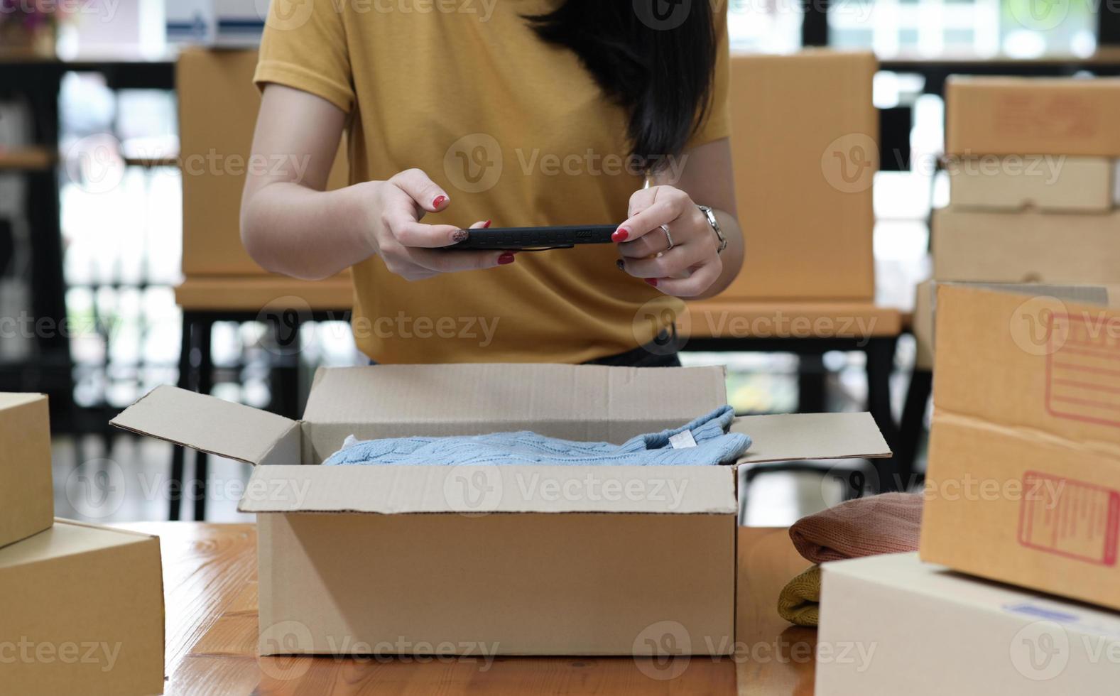 une femme vendant un produit en ligne utilise un smartphone pour prendre une photo du produit dans la boîte.