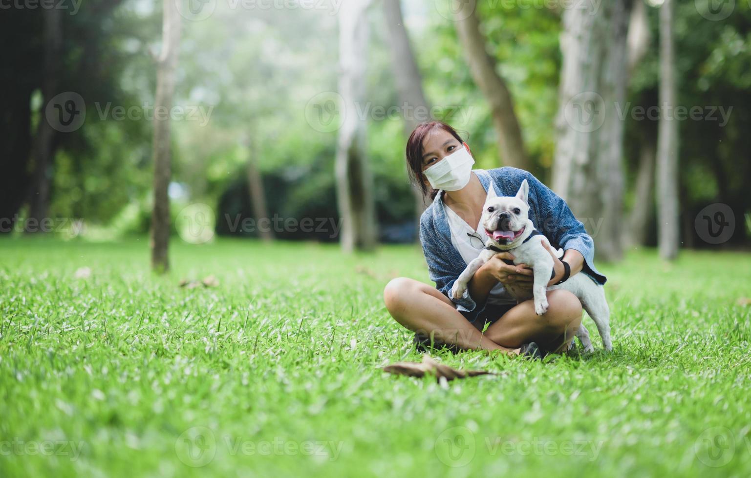 une femme portant un masque médical assise sur la pelouse avec un bouledogue français blanc. photo