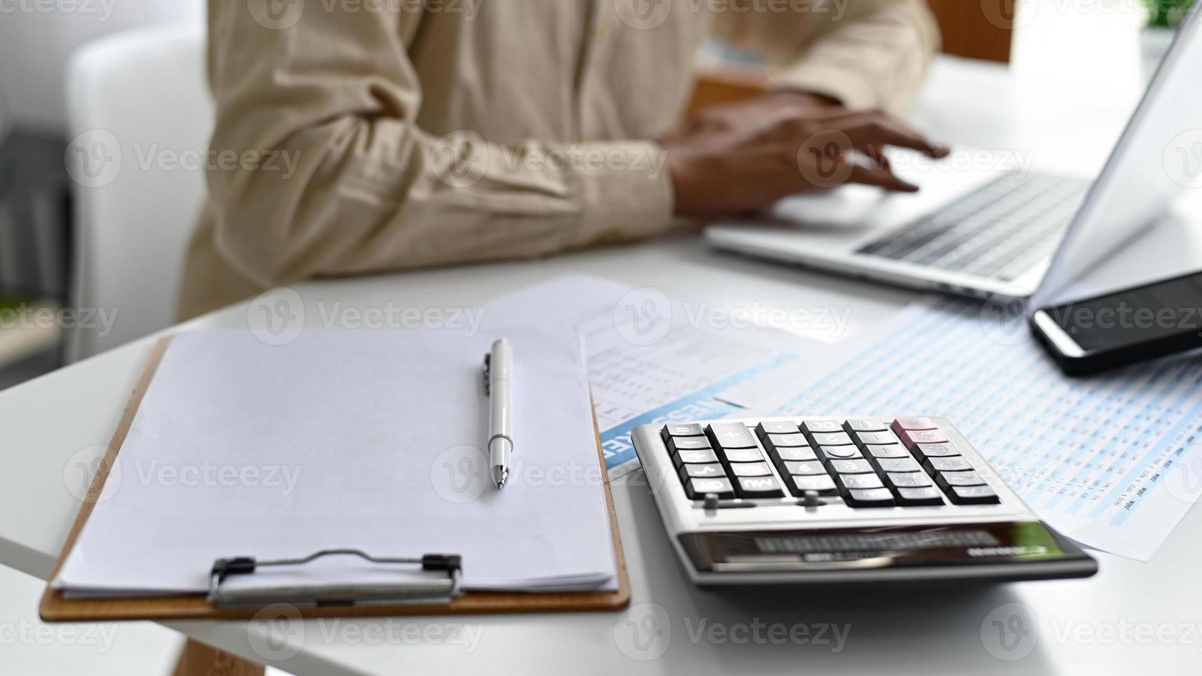 presse-papiers et calculatrice sur le bureau avec des personnes travaillant avec des ordinateurs portables en arrière-plan. photo