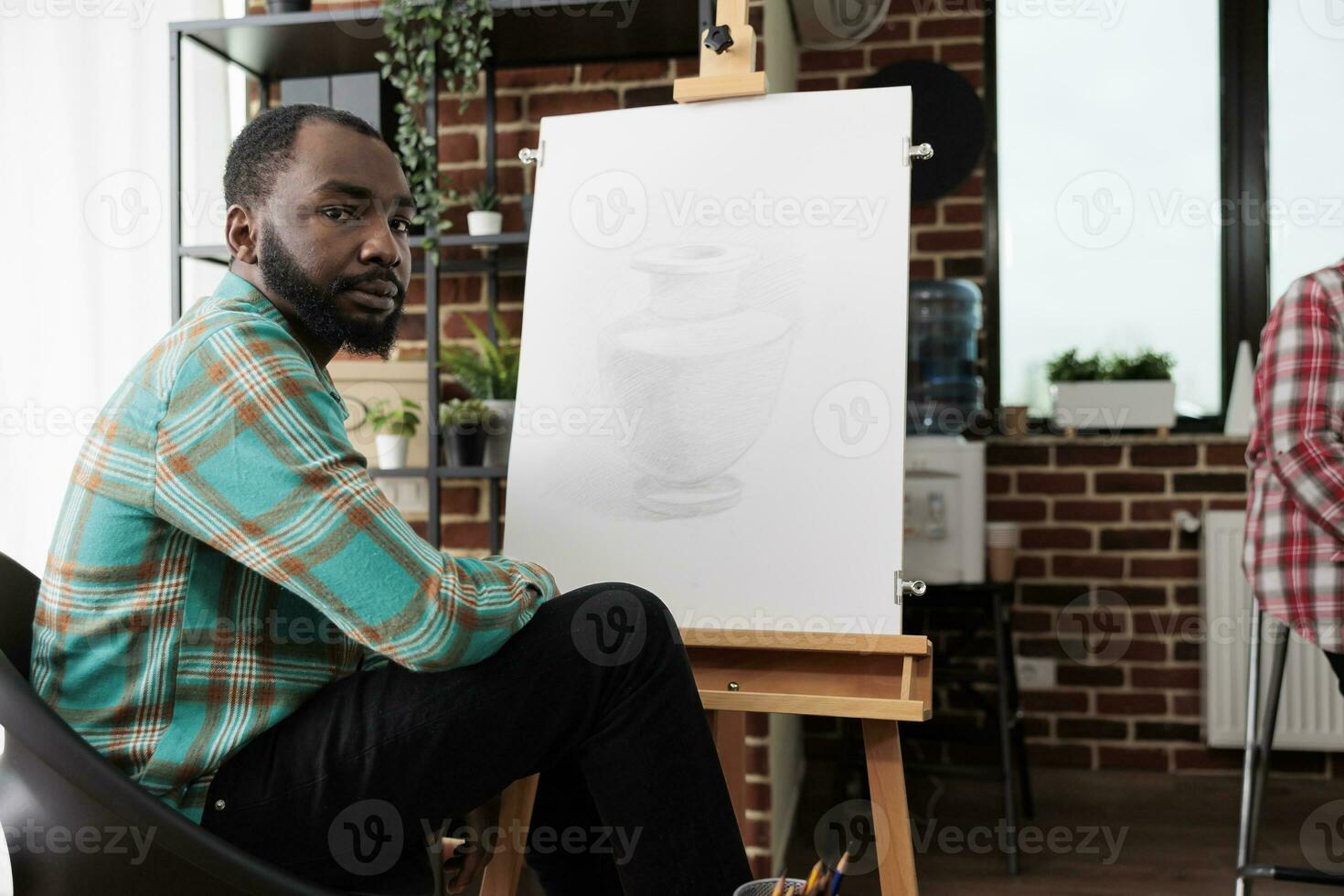 africain américain gars séance à chevalet dans salle de cours et à la recherche à caméra, en train d'étudier bien art. gens participant dans dessin cours de maître, apprentissage divers techniques centré autour crayon croquis. photo