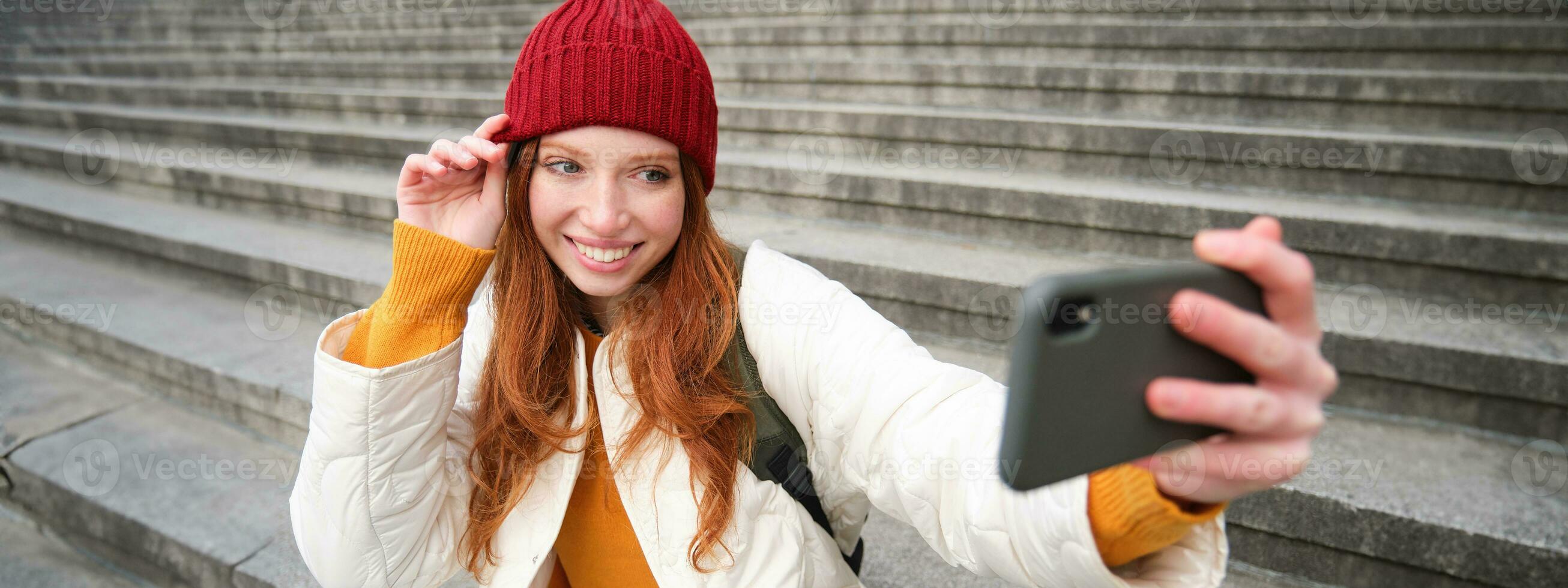 élégant Jeune fille dans rouge chapeau, prend Photos sur téléphone intelligent caméra, fait du selfie comme elle est assis sur escaliers près musée, posant pour photo avec app filtre