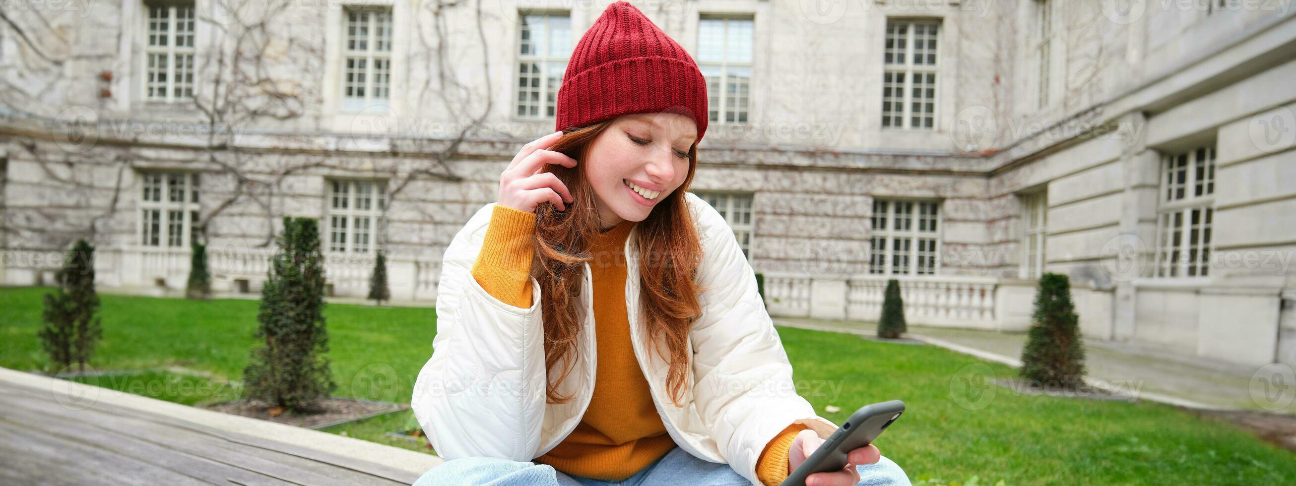 Jeune souriant roux fille est assis sur banc et les usages téléphone intelligent application, lit nouvelles en ligne, montres vidéo sur mobile téléphone tandis que détendu dans parc photo