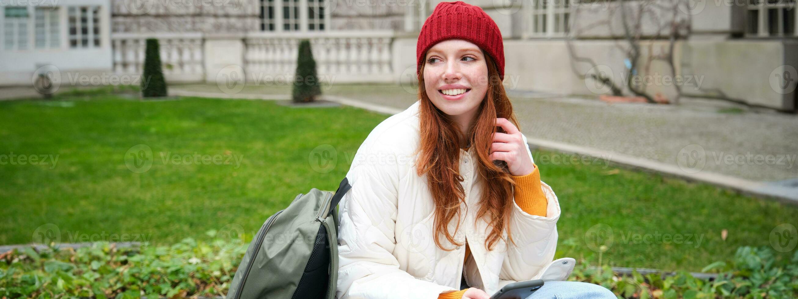 roux fille, femelle étudiant est assis avec mobile téléphone sur banc dans parj, penche sur sa sac à dos. femme navigation social médias app alimentation sur sa téléphone intelligent photo