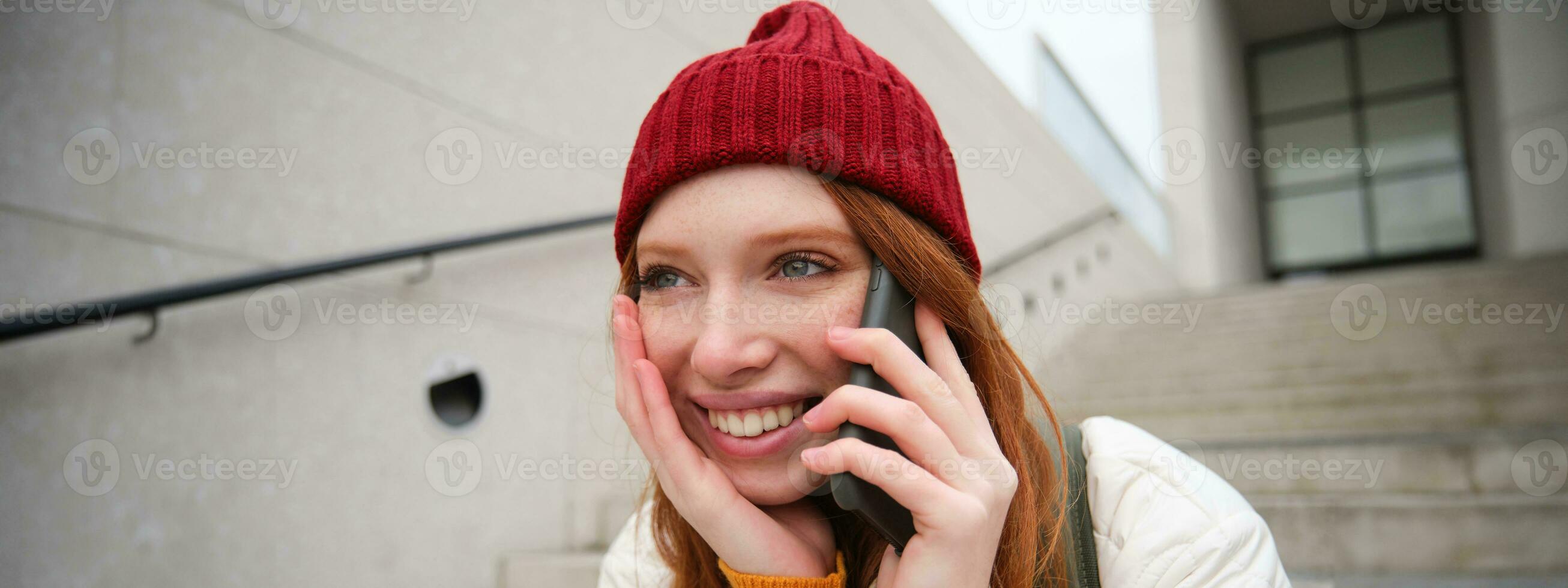 Jeune élégant roux fille dans rouge chapeau, est assis sur rue et pourparlers sur mobile téléphone, a Téléphone conversation, anneaux sa ami tandis que se détend en plein air photo