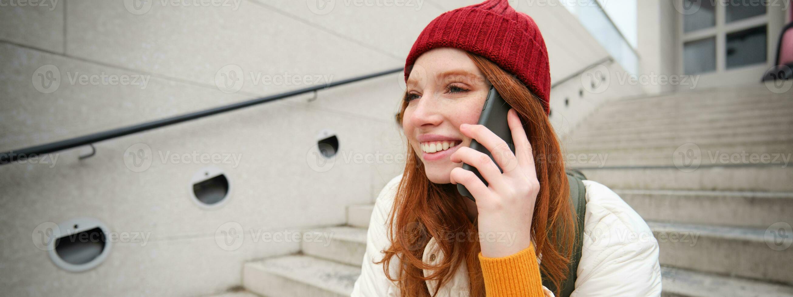 Jeune élégant roux fille dans rouge chapeau, est assis sur rue et pourparlers sur mobile téléphone, a Téléphone conversation, anneaux sa ami tandis que se détend en plein air photo