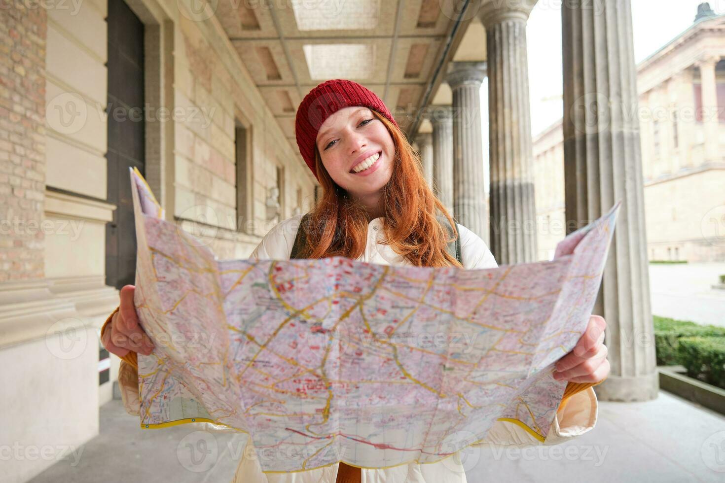 aventureux roux fille des promenades dans ville avec papier carte, explore ville comme touristique, regards pour populaire tourisme attractions, regards autour excité et sourit photo