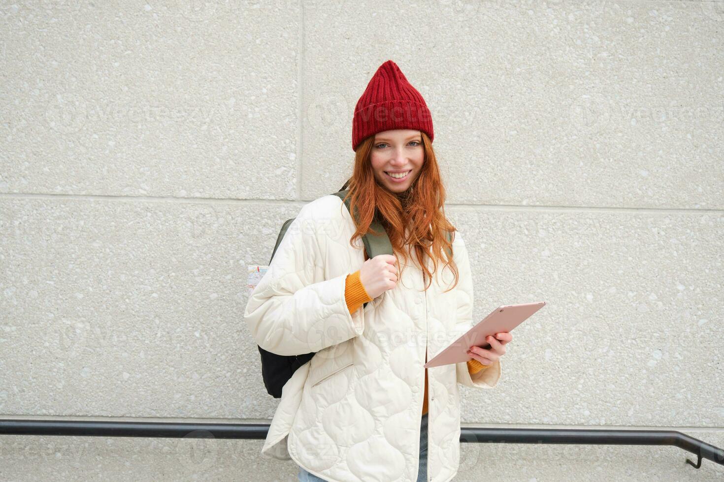 Jeune roux femme avec rouge chapeau, les usages sa numérique tablette en plein air, des stands sur rue avec gadget, connecte à Wifi l'Internet et recherches pour une emplacement dans l'Internet photo