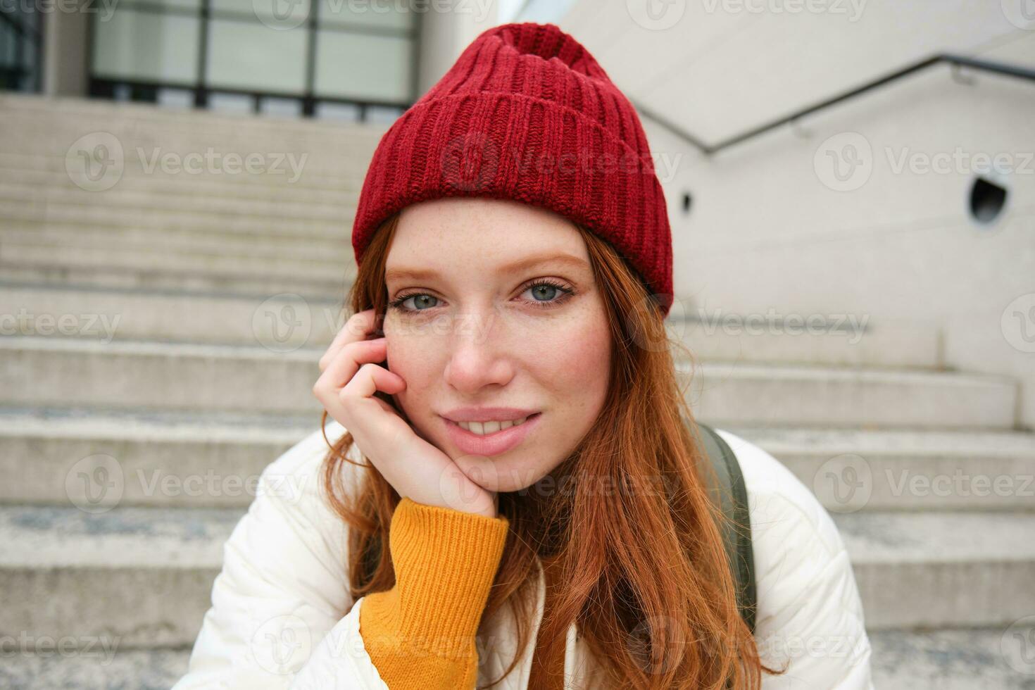 proche en haut portrait de magnifique roux fille dans rouge chapeau, Urbain femme avec taches de rousseur et gingembre cheveux, est assis sur escaliers sur rue, sourit et regards magnifique photo